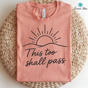 May include: A coral-colored t-shirt with a black graphic of a sun rising over a wavy line. The text "This too shall pass" is written in a cursive font below the graphic. The shirt is folded on a woven mat.