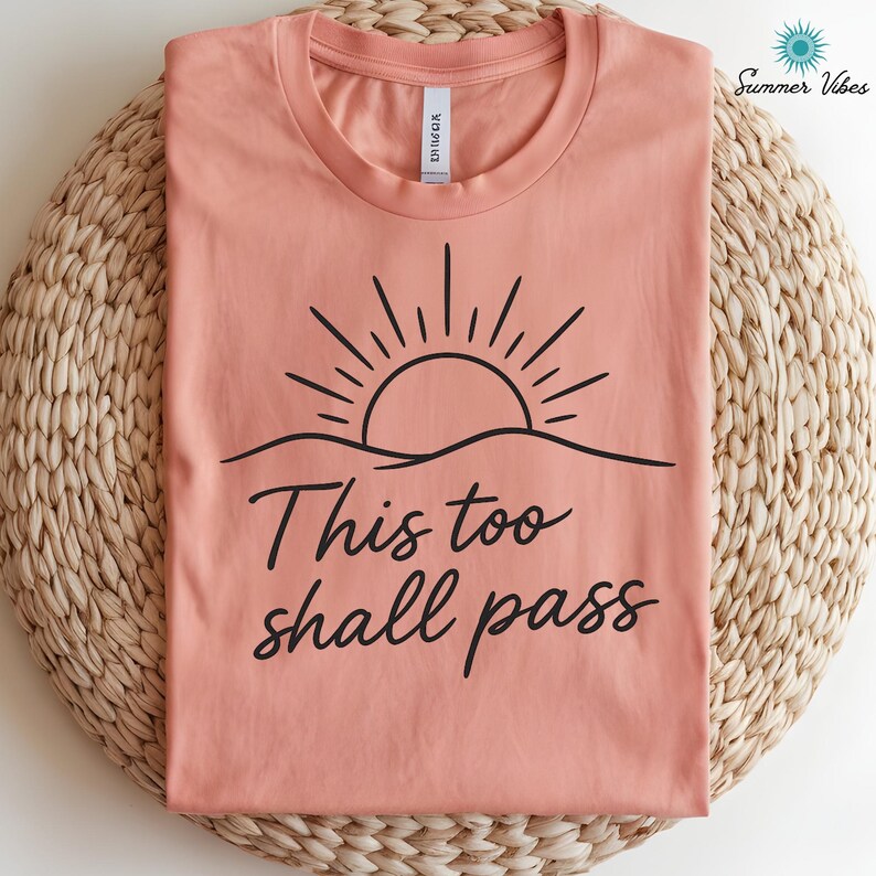 May include: A coral-colored t-shirt with a black graphic of a sun rising over a wavy line. The text "This too shall pass" is written in a cursive font below the graphic. The shirt is folded on a woven mat.