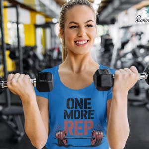 May include: A woman wearing a blue t-shirt with the text "ONE MORE REP" in black and red, holding two black dumbbells. The background is a gym with exercise equipment.
