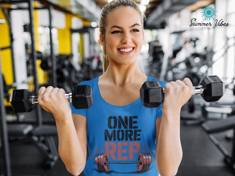 May include: A woman wearing a blue t-shirt with the text "ONE MORE REP" in black and red, holding two black dumbbells. The background is a gym with exercise equipment.