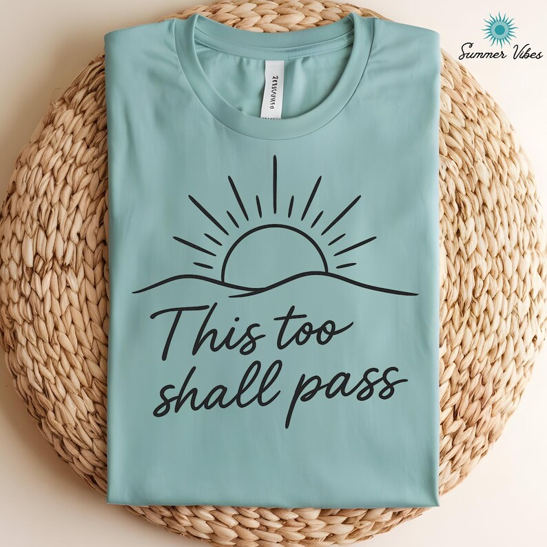 May include: A light teal t-shirt with a black sun and wave design, and the words "This too shall pass" in a script font. The shirt is folded on a woven, light brown surface. The words "Summer Vibes" are in the upper right corner.
