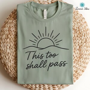 May include: Sage green t-shirt with a black graphic of a sun rising over a wavy horizon. The words "This too shall pass" are written in a cursive font below the graphic. The shirt is displayed on a woven, light brown surface.