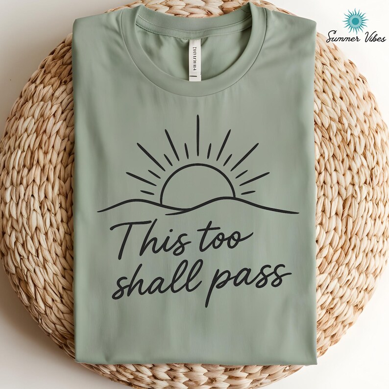 May include: Sage green t-shirt with a black graphic of a sun rising over a wavy horizon. The words "This too shall pass" are written in a cursive font below the graphic. The shirt is displayed on a woven, light brown surface.
