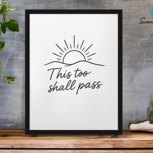 May include: Black framed art print with a minimalist design. The artwork features a sun rising over a wavy line, with the words "This too shall pass" in a handwritten font. The background is white, and the print is displayed on a wooden surface.