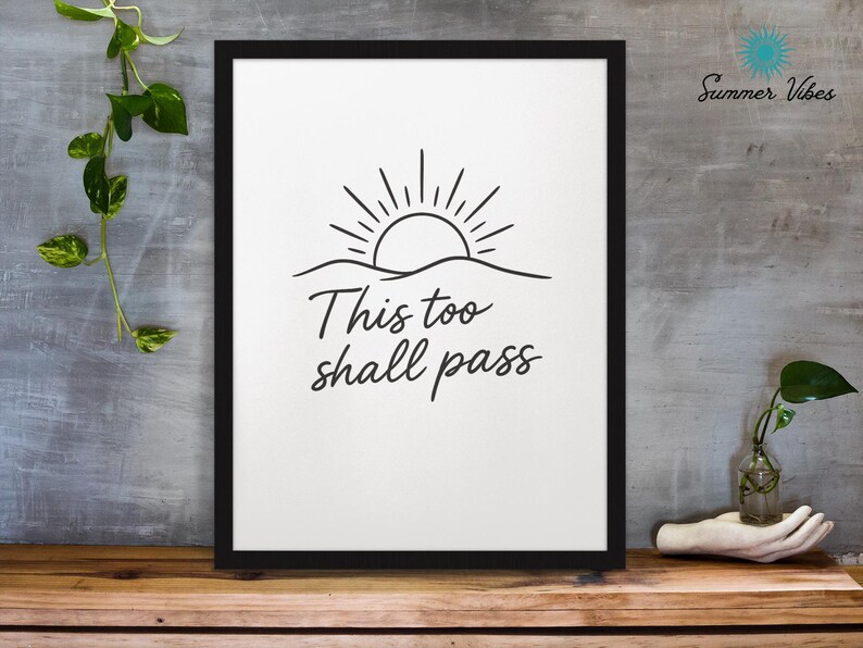 May include: Black framed art print with a minimalist design. The artwork features a sun rising over a wavy line, with the words "This too shall pass" in a handwritten font. The background is white, and the print is displayed on a wooden surface.