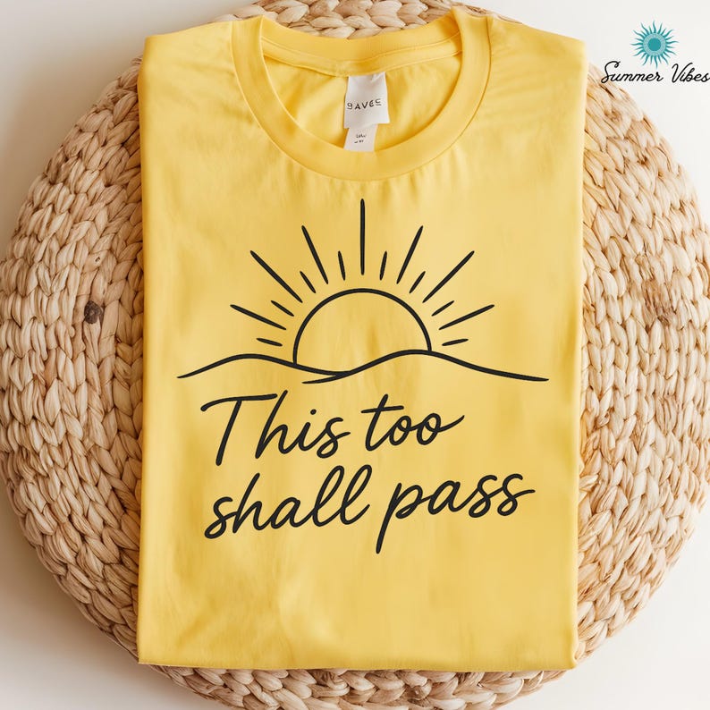 May include: Yellow t-shirt with a black graphic of a sun rising over a horizon and the phrase "This too shall pass" in a cursive font. The shirt is folded on a woven mat. The words "Summer Vibes" are in the upper right corner.