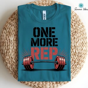 May include: Teal t-shirt with the text "ONE MORE REP" in black and red, featuring a barbell graphic. The shirt has a crew neck and is folded on a woven surface. The words "Summer Vibes" are in the upper right corner.