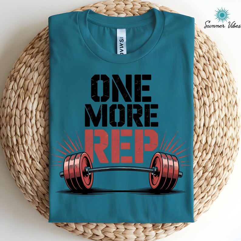 May include: Teal t-shirt with the text "ONE MORE REP" in black and red, featuring a barbell graphic. The shirt has a crew neck and is folded on a woven surface. The words "Summer Vibes" are in the upper right corner.