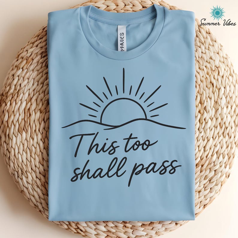 May include: A light blue t-shirt with a black graphic of a sun rising over a wavy line. The text "This too shall pass" is below the graphic. The shirt is folded on a woven surface, with "Summer Vibes" text and a sun logo in the upper right corner.