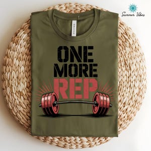 May include: Olive green t-shirt with the text "ONE MORE REP" in black and red. A barbell graphic is centered below the text. The shirt is folded on a woven, circular surface. The words "Summer Vibes" are in the upper right corner.