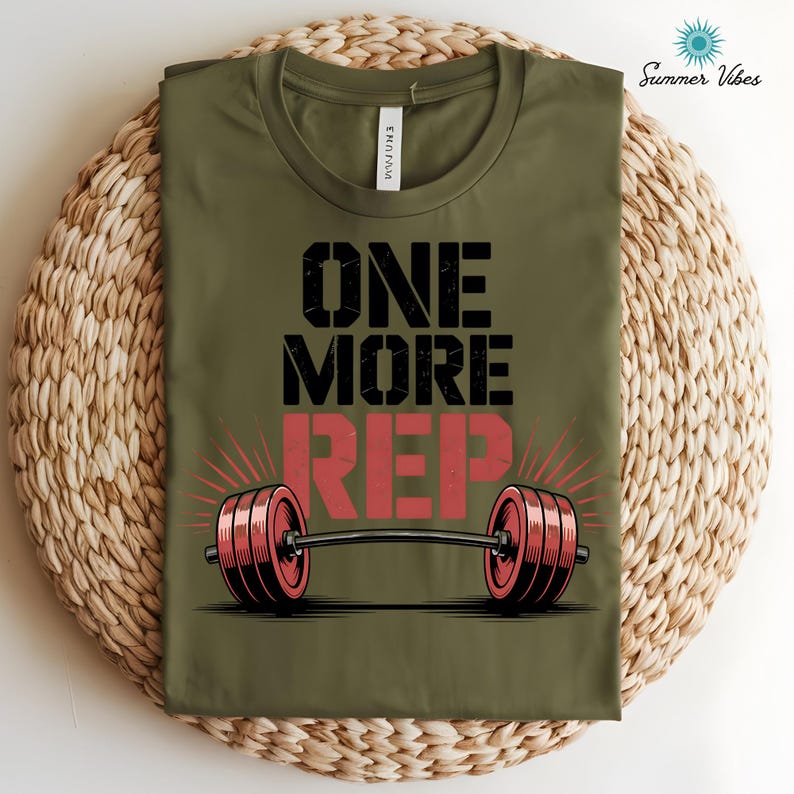 May include: Olive green t-shirt with the text "ONE MORE REP" in black and red. A barbell graphic is centered below the text. The shirt is folded on a woven, circular surface. The words "Summer Vibes" are in the upper right corner.