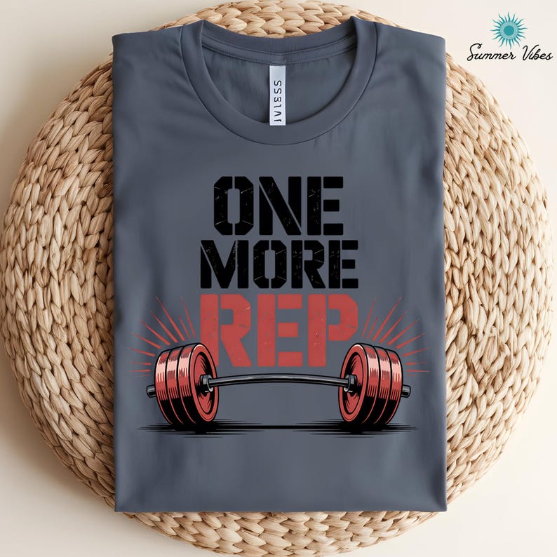 May include: A gray t-shirt with the text "ONE MORE REP" in black and red, featuring a barbell graphic. The shirt has a crew neck and is made of a soft material. The design is centered on the front of the shirt.