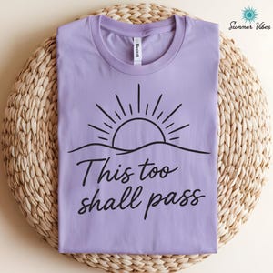 May include: A lilac-colored t-shirt with a black graphic of a sun rising over a wavy line. The text "This too shall pass" is written in a cursive font below the graphic. The shirt is folded neatly on a woven surface.