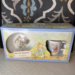 Puede incluir: Un juego de cuenco y taza de cerámica Peter Rabbit by Wedgwood en una caja de presentación rectangular de cartón. La caja presenta ilustraciones de Peter Rabbit en un jardín, con el texto Peter Rabbit by Wedgwood impreso en el frente.