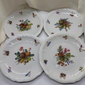 Puede incluir: Juego de cuatro platos de postre de porcelana blanca con un diseño floral y de mariposas. Cada plato presenta un arreglo floral diferente con piñas, bayas y hojas. Los platos tienen un borde festoneado.