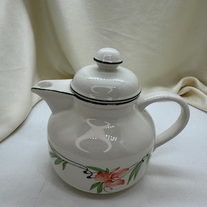 Puede incluir: Tetera de cerámica blanca con tapa, asa y boquilla. Decorada con un diseño floral en rosa y verde. La tapa tiene un pomo verde y ribete negro. La tetera está sobre una superficie blanca.