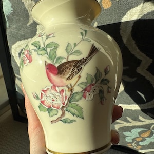 May include: A cream-colored ceramic vase featuring a painted illustration of a small bird with a pink head and brown wings perched on a flowering branch with pink and white blossoms. The vase has a gold-trimmed rim and a gold-trimmed base.