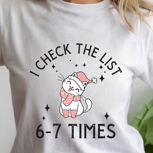 Könnte beinhalten: Weißes T-Shirt mit einer Cartoon-Katze, die einen Weihnachtsmannhut und einen Schal trägt. Der Text "I CHECK THE LIST" wölbt sich über der Katze, darunter steht "6-7 TIMES". Die Katze ist weiß mit rosa Akzenten.