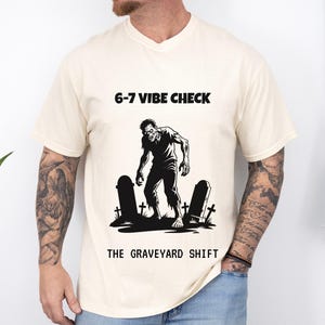 Könnte beinhalten: Cremefarbenes T-Shirt mit einer schwarz-weißen Zombie-Grafik, die auf einem Friedhof steht. Der Text "6-7 VIBE CHECK" steht über der Grafik, und "THE GRAVEYARD SHIFT" steht darunter.