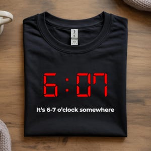Könnte beinhalten: Schwarzes T-Shirt mit roter Digitaluhr-Anzeige, die 6:07 anzeigt. Darunter steht der Text "It's 6-7 o'clock somewhere". Das T-Shirt liegt gefaltet auf einer Holzoberfläche.