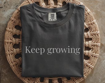 May include: A dark gray t-shirt with the words "Keep growing" printed in a light gray font. The shirt is folded and placed on a round, woven wicker placemat. The shirt has a crew neck and short sleeves.