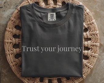 May include: A dark gray t-shirt with the phrase "Trust your journey" printed in white. The shirt is displayed on a round, woven wicker placemat. The shirt has a crew neck and short sleeves.