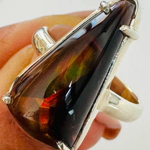 May include: A silver ring featuring a large, triangular-shaped ammolite gemstone. The stone displays a dark, polished surface with iridescent flashes of deep red, orange, and green colors. The gem is secured in a simple silver prong setting.