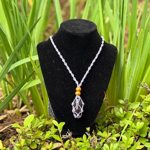 Può includere: Una collana fatta a mano con un ciondolo di cristallo scuro in un macramè intrecciato viola chiaro. La collana ha un cordino viola chiaro con due perline di legno. Il ciondolo è esposto su un manichino nero su uno sfondo di fogliame verde.
