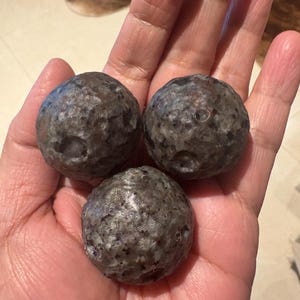 May include: Three dark gray, textured spheres are held in a hand. The spheres have a rough, pitted surface and appear to be made of a metallic material. The overall impression is of a collection of unique, handcrafted objects.