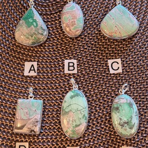 May include: Six silver pendant necklaces with natural stone pendants. The pendants have a mix of green, tan, and white colors. The pendants are in various shapes, including teardrop, oval, and rectangular.
