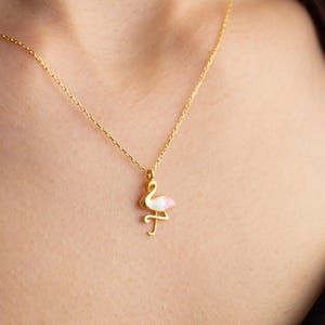 May include: A gold-colored necklace featuring a flamingo pendant with pink and white enamel detailing. The flamingo is depicted in a stylized pose, suspended from a delicate chain. The necklace is worn around the neck.