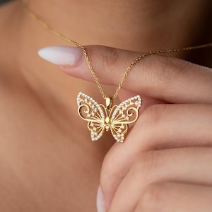May include: A gold-colored butterfly pendant necklace. The butterfly design features intricate filigree detailing and is embellished with small, sparkling gemstones. The necklace chain is also gold-colored.
