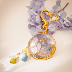 Puede incluir: Un llavero dorado con un colgante redondo lleno de flores azules y blancas secas y copos de oro. El llavero tiene un cierre en forma de corazón y una cadena con cuentas azules, verdes y blancas.