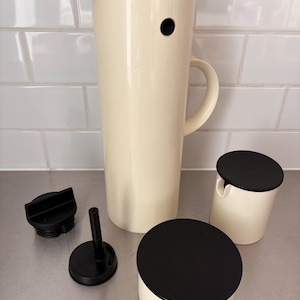 Puede incluir: Un servidor de bebidas de color crema con tapa y asa negras, acompañado de dos recipientes de color crema con tapas negras y un tapón negro. El servidor tiene una forma alta y cilíndrica. Los recipientes son redondos y cilíndricos.