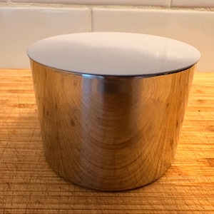 Può includere: Un contenitore cilindrico con una finitura metallica lucida e un coperchio bianco e arrotondato. Il contenitore è posto su una superficie di legno con venature e graffi visibili. Il design è semplice e moderno.