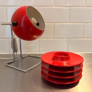 May include: A red, mid-century modern desk lamp with a spherical shade and a stack of red plates. The lamp has a chrome base and a black switch. The plates are stacked on top of each other.