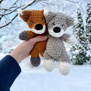 May include: Two crocheted stuffed animals: a brown fox and a gray wolf. The fox has a white muzzle and ears, with a black nose. The wolf has a white muzzle, ears, and paws, with a black nose. Both are held up against a snowy background.