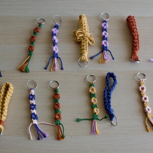 Może przedstawiać: Kolekcja ręcznie robionych breloków z makramy w różnych kolorach, w tym niebieskim, zielonym, żółtym, pomarańczowym i fioletowym. Niektóre breloki mają kwiatowe wzory, inne skomplikowane wzory tkane. Każdy brelok ma metalowy pierścień.