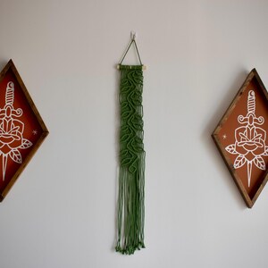 May include: Wall decor set featuring two diamond-shaped wooden plaques with a dagger and rose design in white on an orange background. A green macrame wall hanging is centered between the plaques.