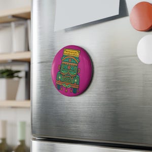 May include: A round, magenta magnet featuring a colorful illustration of a decorated truck. The truck is adorned with intricate patterns in green, orange, and blue. A small, rectangular sign with text is at the top of the truck.