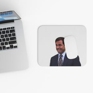 以下が含まれることがあります： 驚いた表情のスーツ姿の男性の画像がプリントされた白いマウスパッド。白いコンピューターマウスがマウスパッドの右側にあります。ラップトップが左上隅に見えます。