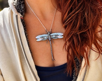 May include: A silver-toned dragonfly pendant necklace featuring iridescent light blue wings and a body encrusted with small dark blue crystals. The dragonfly is suspended from a delicate silver chain that rests against the chest area.