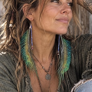 May include: Dangle earrings featuring long, iridescent green and gold peacock feathers attached to thin, vertical blue crystal-studded bars. Each earring terminates in a small, round metallic bead at the base of the blue crystal chain.