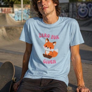 May include: A light blue short-sleeve t-shirt featuring a graphic of a small orange fox with a white-tipped tail and pink cheeks. Above and below the fox, the text reads "ZERO FOX GIVEN" in a rounded, pink, bubble-style font.
