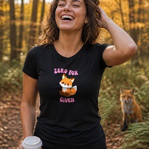 May include: A black short-sleeved t-shirt featuring a graphic of a small orange and white fox sitting down. Above the fox is the text "ZERO FOX" and below the fox is the text "GIVEN" in pink lettering. The shirt has a round neckline and a fitted cut.