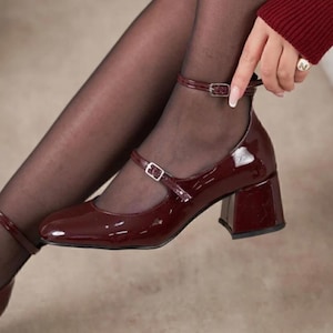 May include: Burgundy patent leather Mary Jane style shoes with a block heel and ankle strap. The shoes have a glossy finish and a square toe. The ankle strap features a small silver buckle. The shoes are worn with black tights.