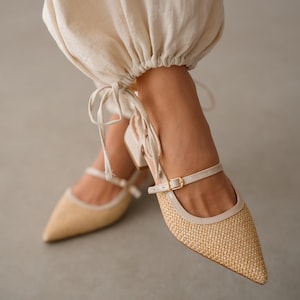 May include: Close-up of beige raffia-textured shoes with pointed toes and ankle straps. The shoes have a small block heel and a cream-coloured trim. The straps are secured with small gold buckles.