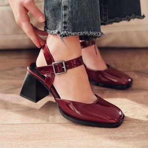 May include: Burgundy-colored, glossy heeled sandals with a square toe and a black, block heel. The ankle strap features a silver buckle. The shoes are worn with distressed denim jeans.