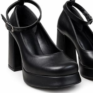 May include: A pair of black platform pumps featuring a square toe, a high block heel, and a thin ankle strap with a silver-toned metal buckle. The shoes have a smooth finish and a thick platform sole that elevates the front of the foot.