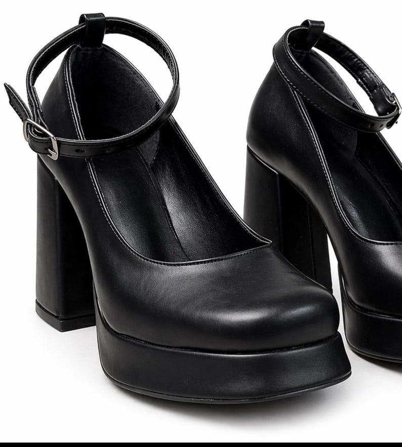 May include: A pair of black platform pumps featuring a square toe, a high block heel, and a thin ankle strap with a silver-toned metal buckle. The shoes have a smooth finish and a thick platform sole that elevates the front of the foot.